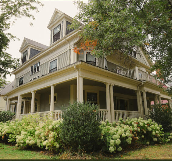 Garden District Napanee two-story white house with dark shutters and a gabled roof sits in a well-kept yard with green grass and vibrant red autumn trees. The symmetrical facade features a central entrance with a small columned portico, and a paved driveway leads up to the house.